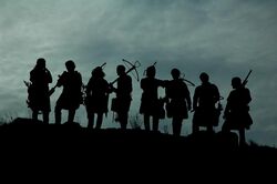 Silhouette of a group of eight medieval style soldiers with crossbows against a cloudy sky.