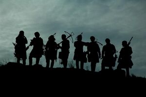 Silhouette of a group of eight medieval style soldiers with crossbows against a cloudy sky.