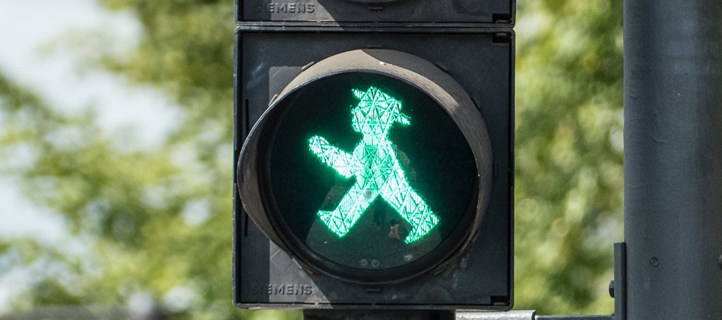 Street light with a green illuminated crossing sign with image of person in hat walking near trees